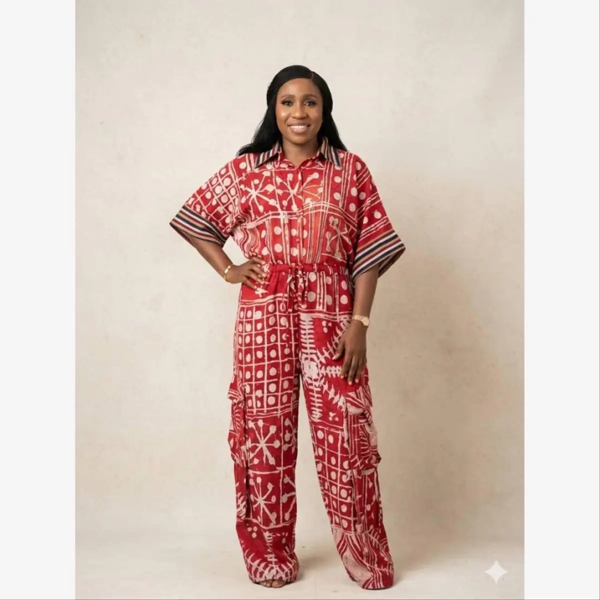 Front-facing full-length view of a woman wearing a handcrafted Adire batik wide-leg cargo jumpsuit in a bold crimson red and cream resist-dyed pattern. The outfit features a collared short-sleeve top with a drawstring waist and cargo pockets on the wide-leg trousers, with multi-stripe sleeve cuff trim. Color: Crimson Red and Cream
