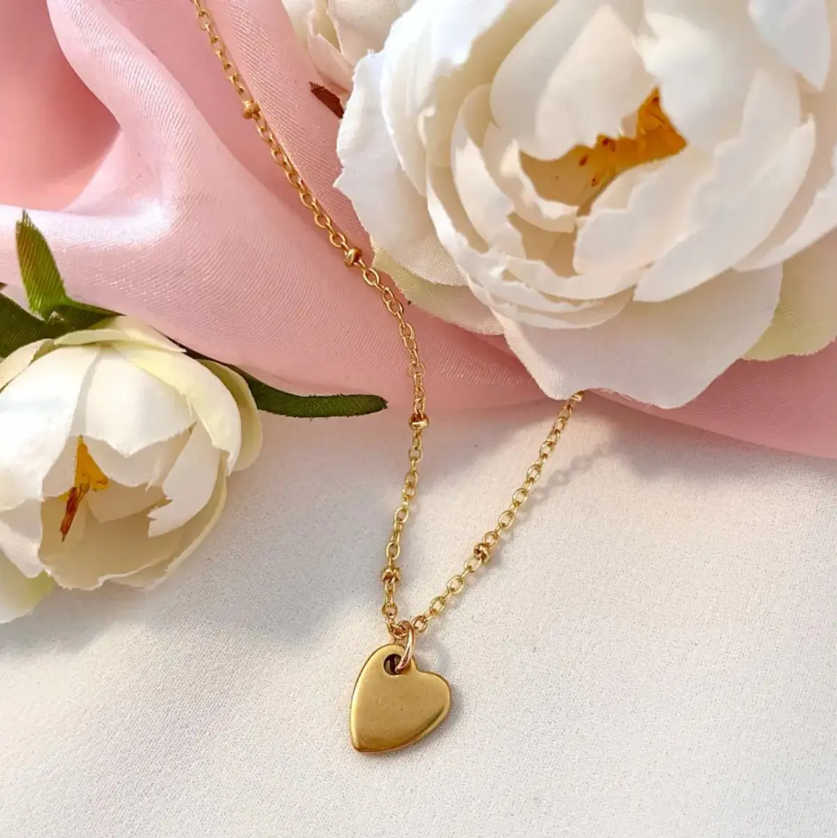 A close-up flat-lay of a handcrafted gold heart pendant necklace resting on a white linen surface, surrounded by soft white peony blooms and a blush pink satin ribbon. The small polished gold heart charm hangs from a delicate satellite chain, glowing warmly against the romantic floral backdrop. Color: Gold