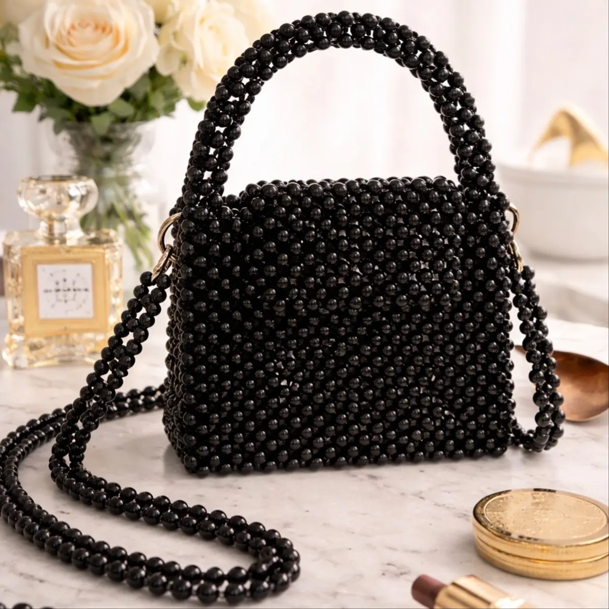 A handcrafted beaded box bag entirely covered in glossy black pearl-finish beads, styled on a marble surface alongside a perfume bottle and gold compact. The bag features a structured silhouette, a beaded top handle, and a long beaded crossbody strap with gold-tone hardware. Color: Black