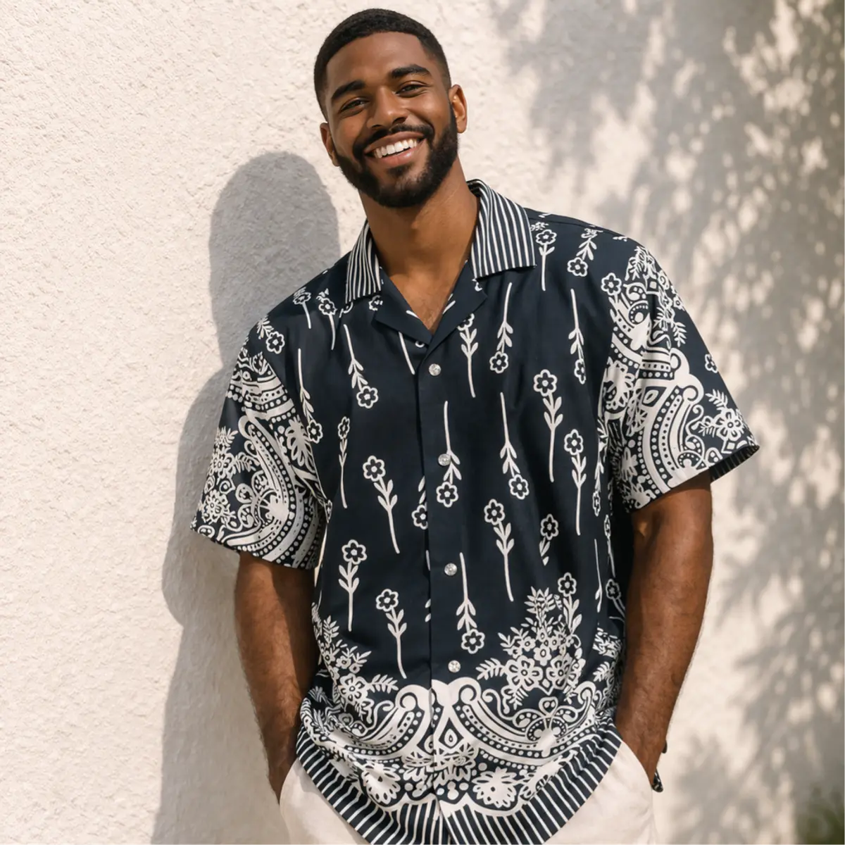 Close-up front view of a man wearing a navy blue and white handcrafted camp collar shirt. The shirt features delicate white floral and botanical stem prints across the body, bold paisley medallion detailing on the sleeves and hem, and a contrast pinstripe collar. Color: Navy Blue and White